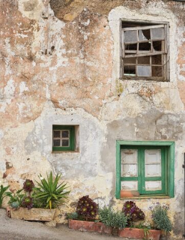 Fachada de una casa de piedra encalada con la pintura descascarillada, una ventana de madera rota en el piso alto y otra pintada de verde abajo, con macetas de bejeques y pitas al pie del muro.