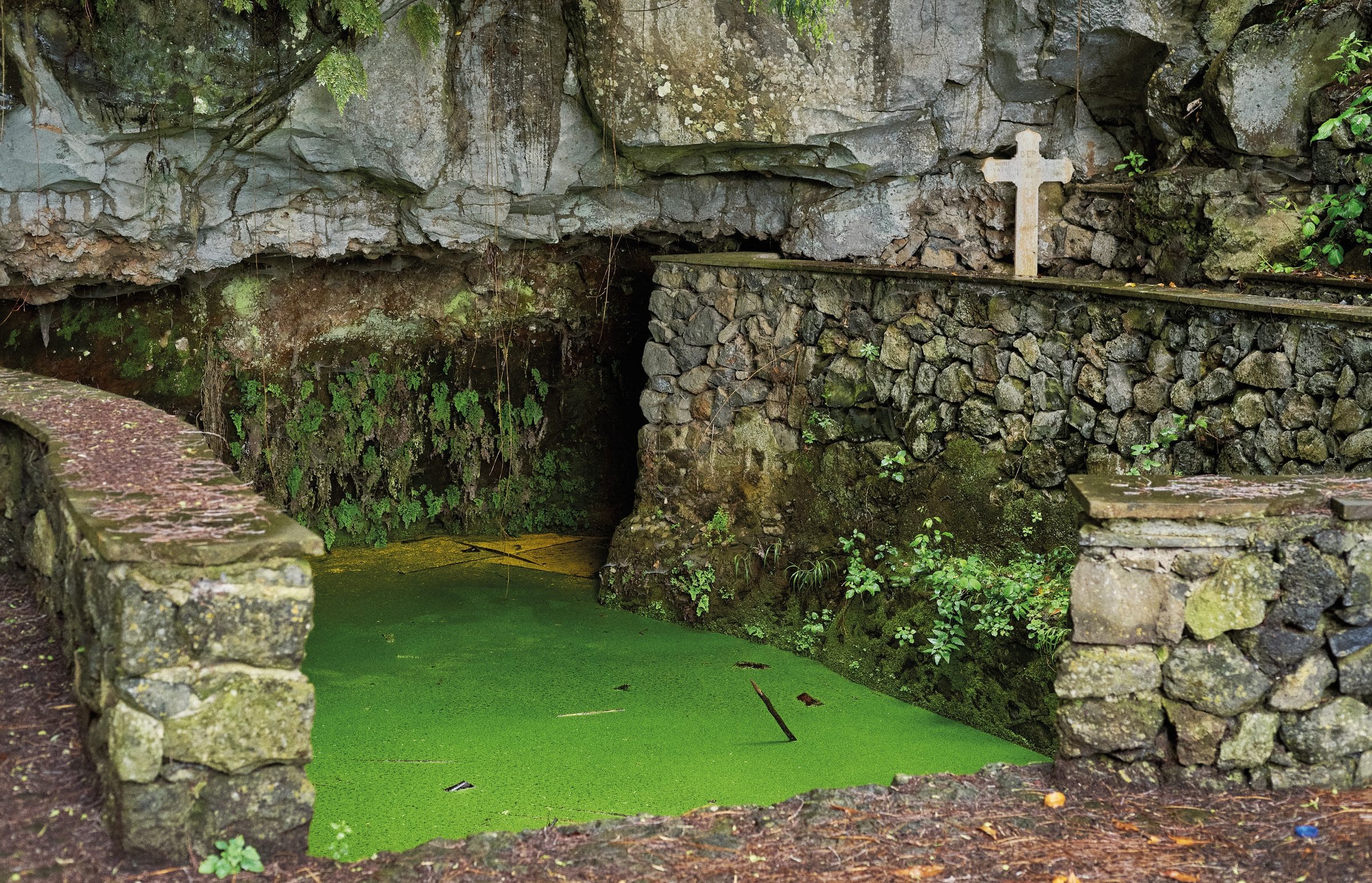 Un lavadero excavado bajo una gran roca, con el agua cubierta por una capa de lenteja de agua verde intenso y una cruz blanca de piedra sobre el muro.