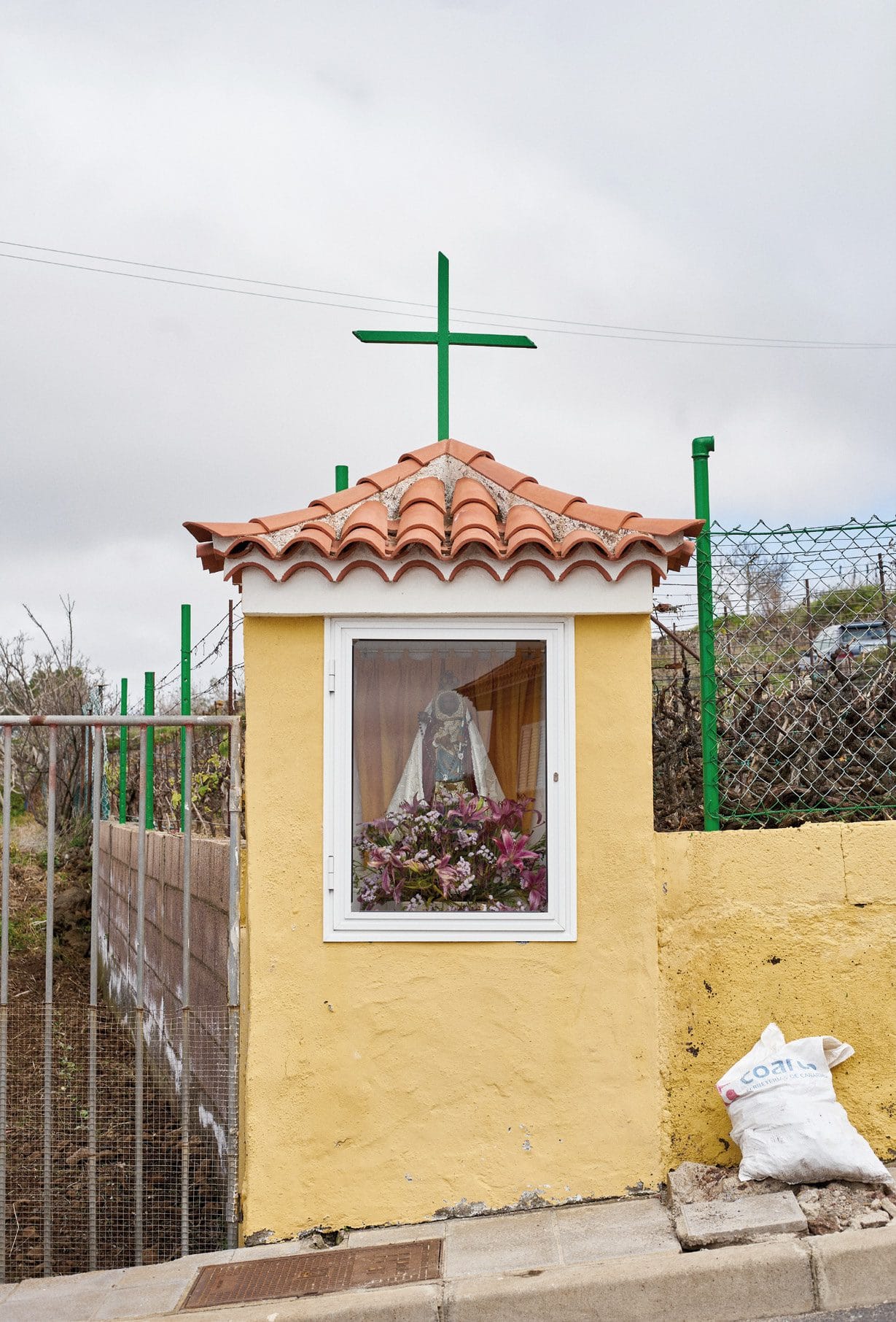 Una ermita en miniatura de fachada amarilla con tejadillo de teja y cruz verde, con una virgen y flores tras el cristal, junto a una valla metálica.