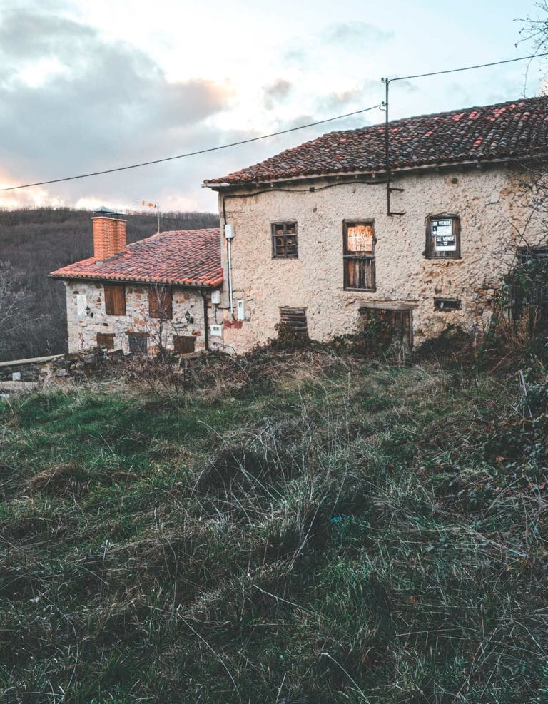 Una casa de piedra abandonada en un pueblo con carteles de «Se vende» en las ventanas, al atardecer.