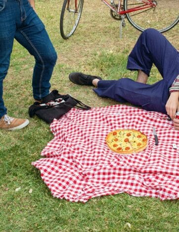 Dos personas de pie y una tercera agachada alrededor de un mantel de cuadros rojos y blancos tendido sobre la hierba, con dos bicicletas apoyadas detrás.