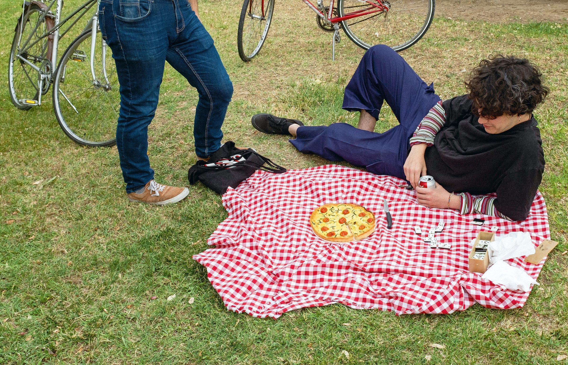 Dos personas de pie y una tercera agachada alrededor de un mantel de cuadros rojos y blancos tendido sobre la hierba, con dos bicicletas apoyadas detrás.