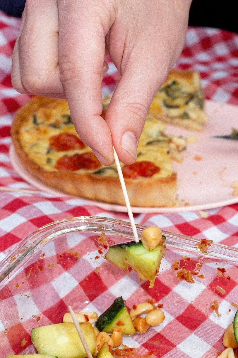 Una mano pincha con un palillo un trozo de pizza casera sobre el mantel de cuadros, junto a un táper con ensalada de patata.