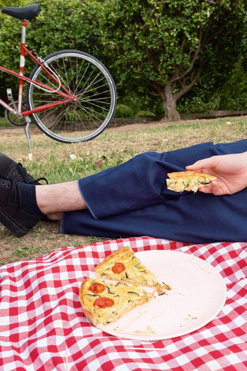 Una persona tumbada en la hierba junto a un plato con un trozo de pizza sobre el mantel de cuadros, con una bicicleta roja al fondo.