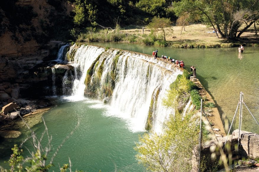 Una cascada ancha con bañistas asomados en el borde superior y en la poza de abajo.