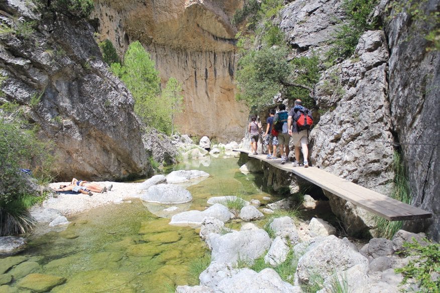 Un grupo de personas cruza en fila una pasarela de madera anclada a la pared de un cañón.