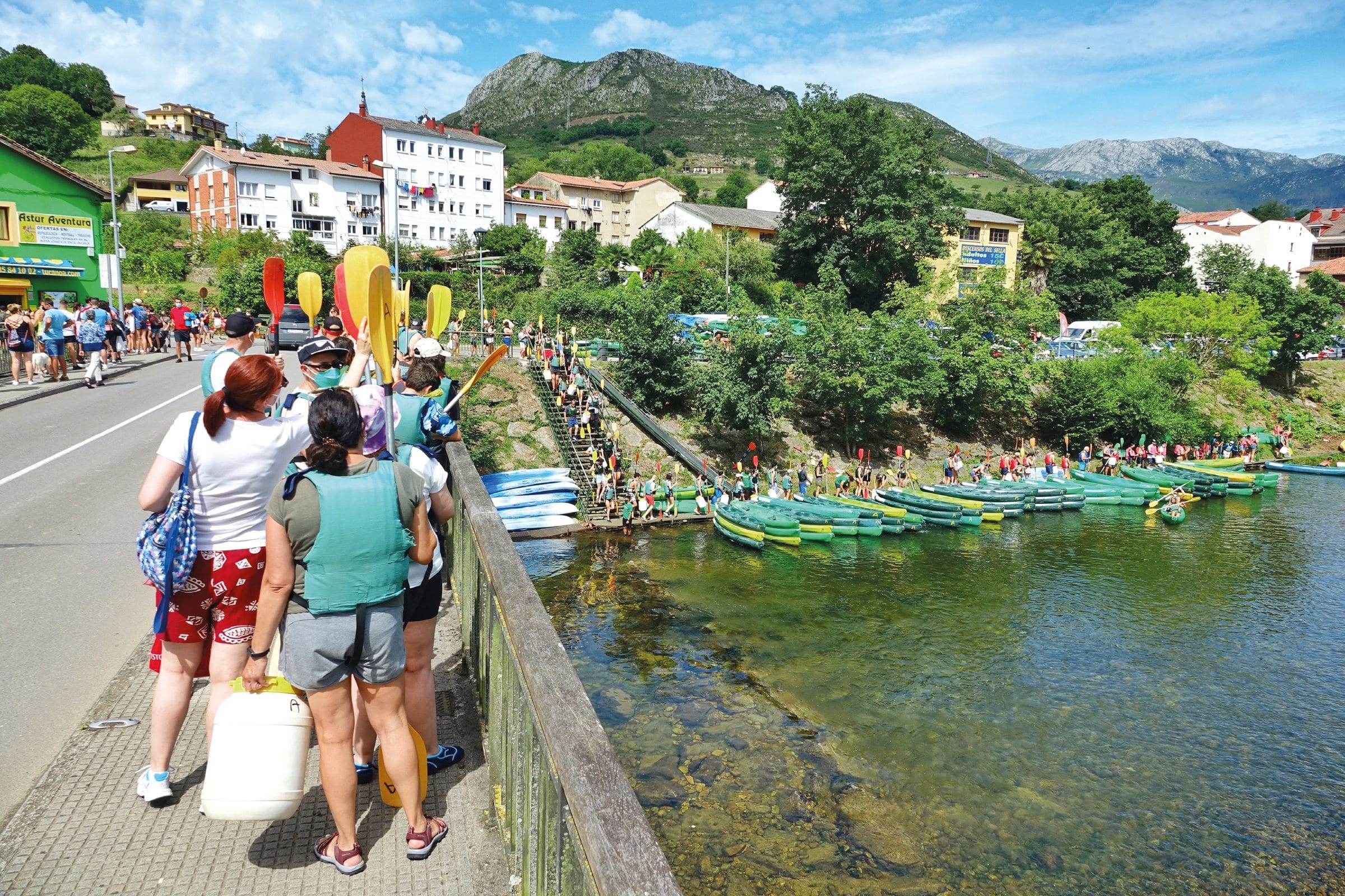 Tres personas de espaldas, apoyadas en una barandilla junto a la carretera, miran un tramo de río lleno de kayaks amarillos.