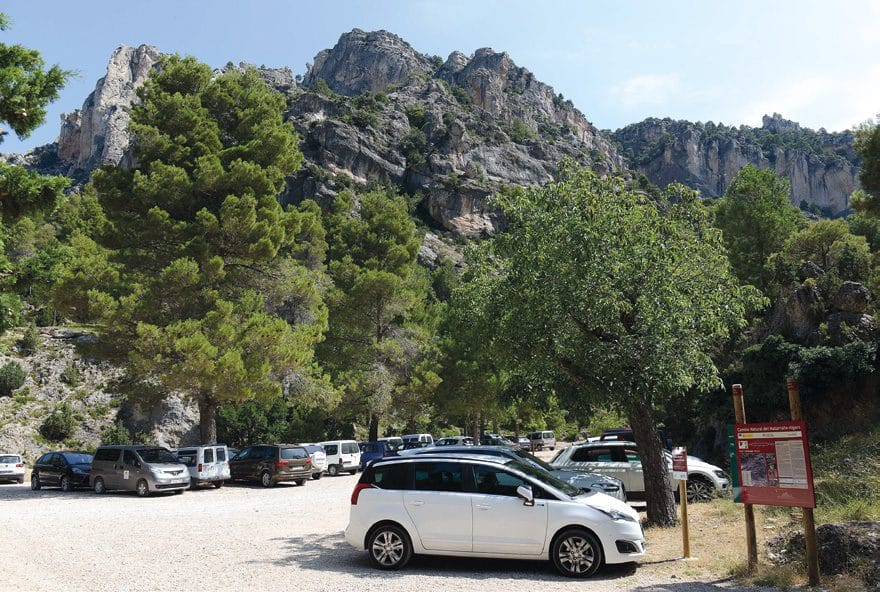 Un aparcamiento de tierra lleno de coches al pie de un farallón rocoso.