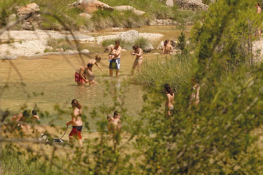Bañistas en una playa fluvial, vistos entre la vegetación de la orilla.